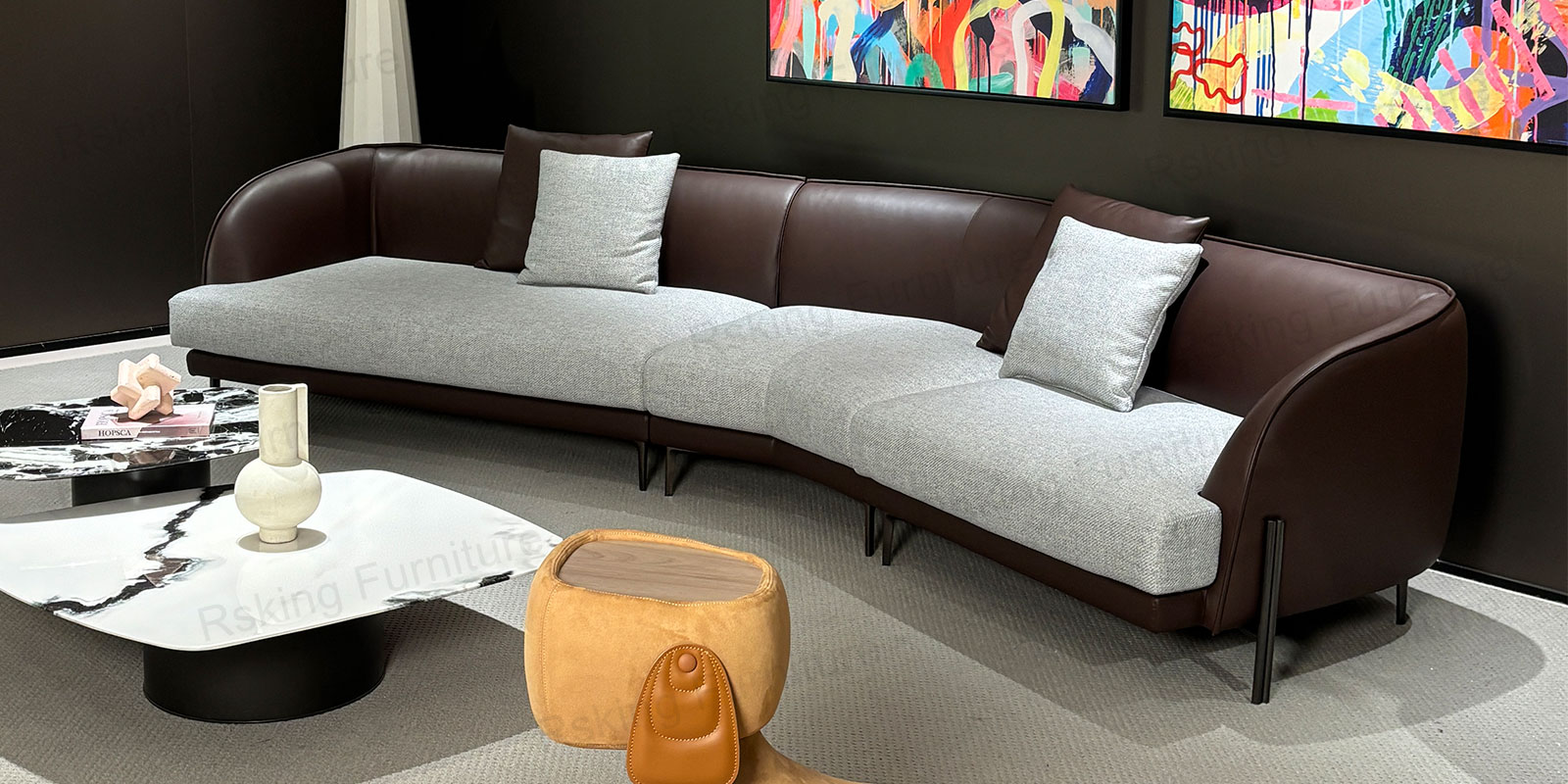 Angled showroom view of the curved leather and fabric sofa beside stone coffee tables and colorful art