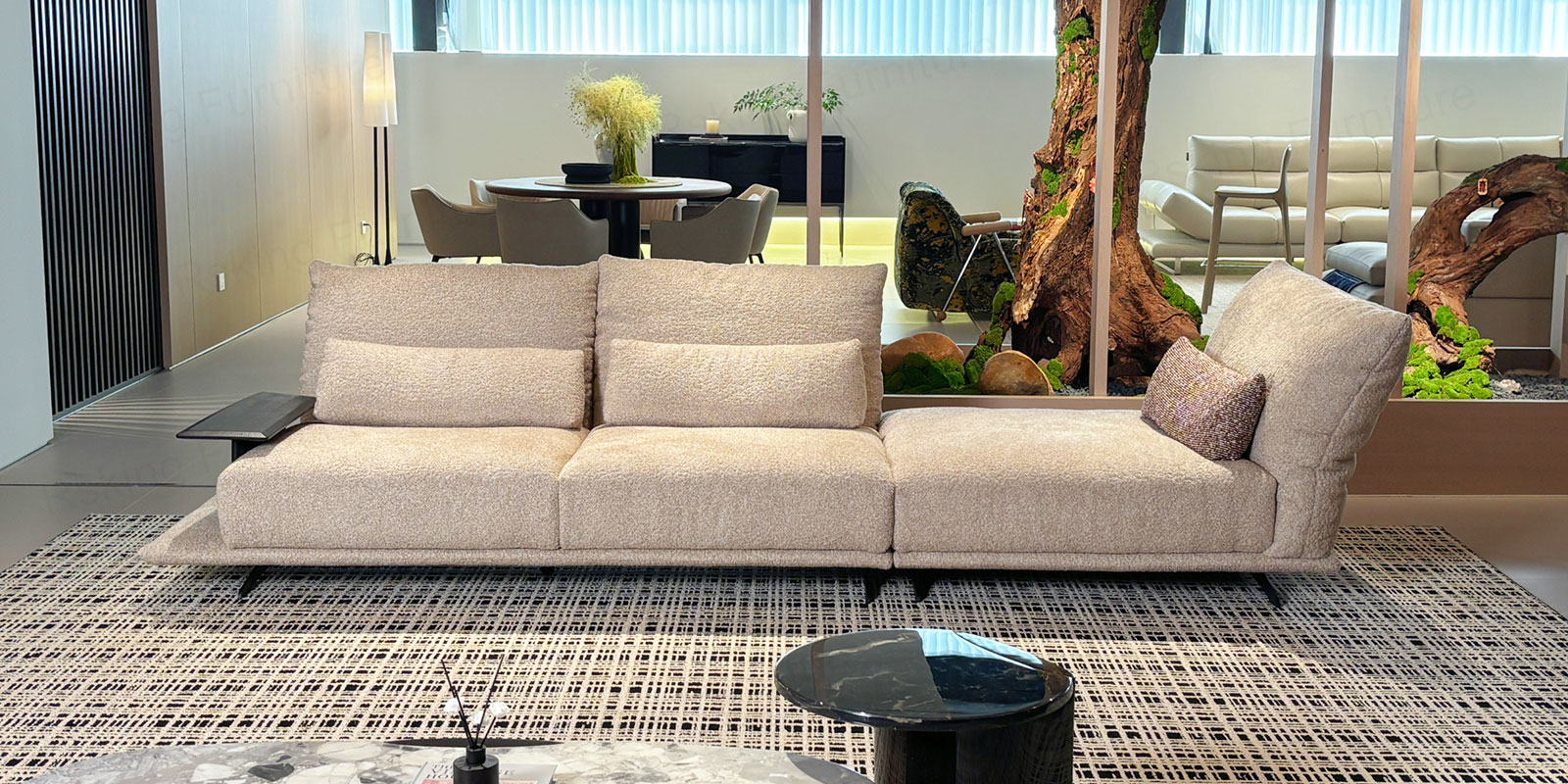 Off-white fabric sofa displayed on a patterned rug in a showroom with natural wood and greenery